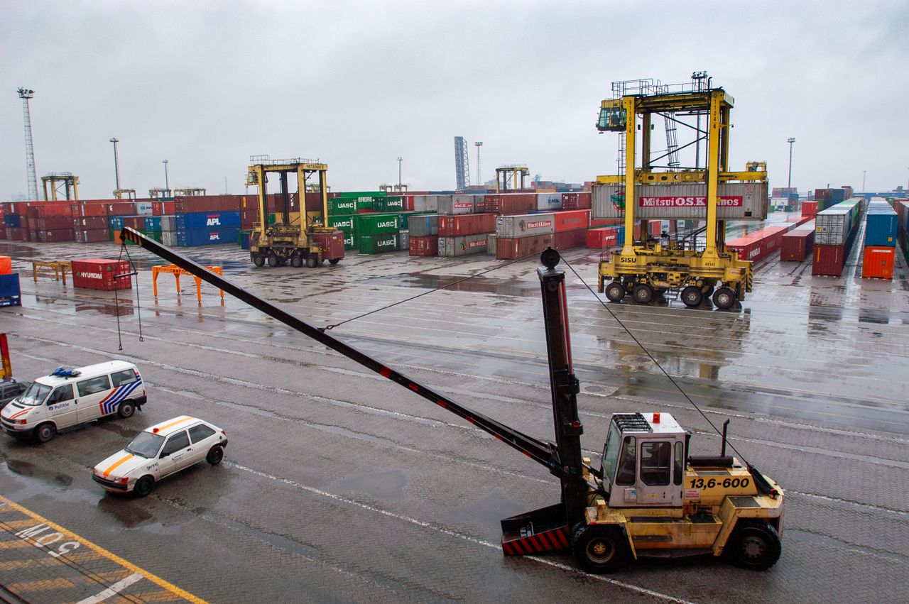 Container terminal with straddle carriers and stacked shipping containers under a cloudy sky.